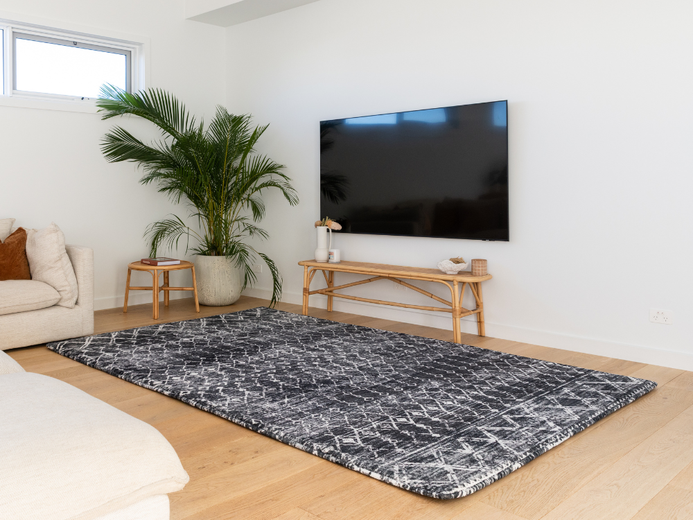 Living room with a large flat-screen TV mounted on the wall, a rug on the floor, and a plant.