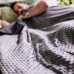 Person lying in bed under a gray blanket with a blurred background