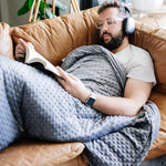 Man lying on a couch with a blanket, reading a book and wearing headphones.