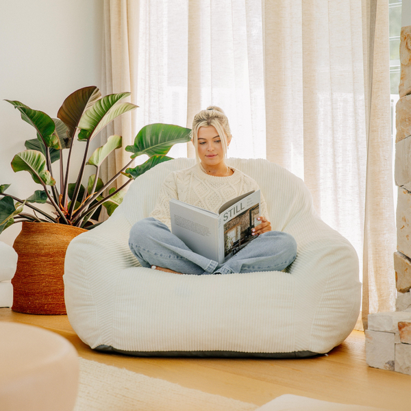 Woman reading a magazine on a large white bean bag chair in a bright room with plants.