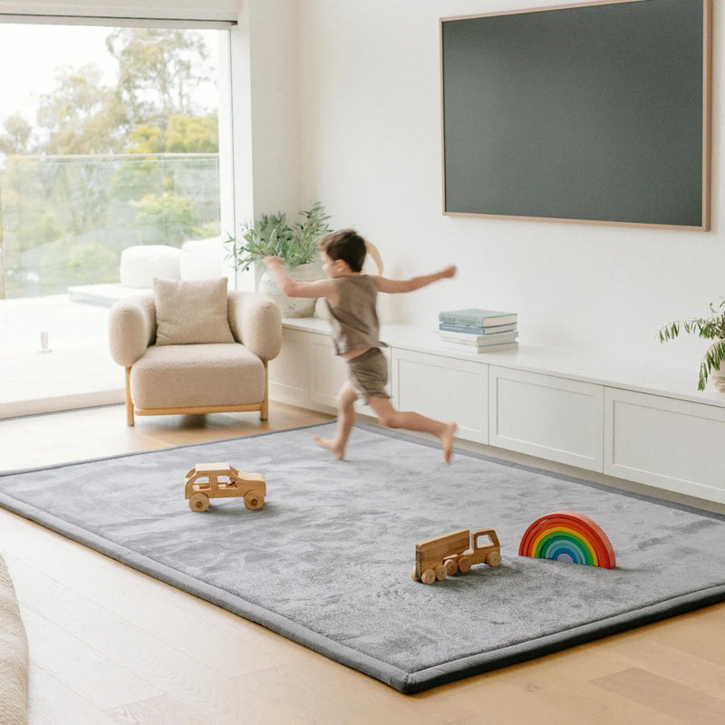 child running on a grey rug wit some small wooder toy cars and a rainbow toy